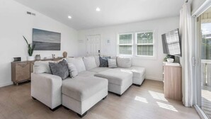 Un salon spacieux comprenant un canapé d’angle, un meuble en bois, un téléviseur à écran plat et une grande fenêtre équipée de stores.