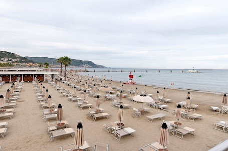 A beach with rows of sun loungers and umbrellas, a pier extending into the sea, and a hill with buildings in the background.. Apartment 'M&M House Beach - 4 Salti Dal Mare' with Mountain View, Wi-Fi and Air Conditioning