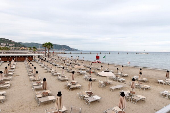 Una playa con filas de tumbonas y sombrillas, un muelle que se adentra en el mar, y una colina con edificios al fondo.