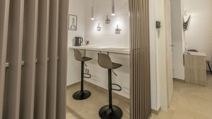 Une cuisine moderne avec un plan de travail blanc, deux tabourets de bar, des luminaires suspendus et une machine à café.