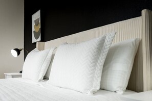 Una cama bien hecha con ropa blanca y almohadas, una lámpara de noche y un cuadro en la pared.