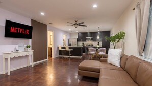 A modern living room with a sofa, a TV mounted on the wall, a ceiling fan, and a kitchen area in the background.