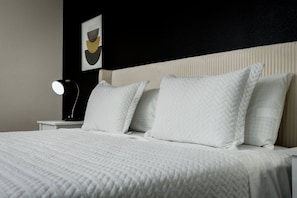 Una cama bien hecha con ropa blanca, cabecera y lámpara de noche.