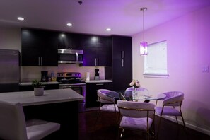 A modern kitchen with dark cabinets, a stainless steel refrigerator, a microwave, and a gas stove. There is a dining area with a glass table and purple chairs.