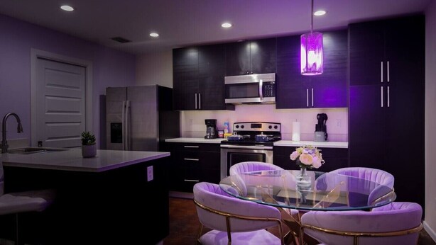 A modern kitchen with dark cabinetry, a central island, and a dining area with purple chairs and a glass table.