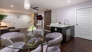 A modern living room with a glass dining table, comfortable seating, a ceiling fan, and a TV mounted on the wall.