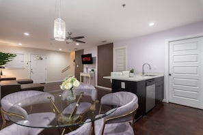 Una sala de estar moderna con una mesa de vidrio, sillas blancas, un ventilador de techo, un televisor y una zona de cocina con un lavamanos y armarios.