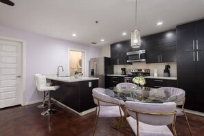 Una cocina moderna con armarios oscuros, electrodomésticos de acero inoxidable, una mesa de comedor de vidrio con sillas blancas y una lámpara colgante.
