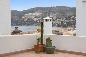 Een balkon met terracotta potten en cactussen, met uitzicht op een kustplaatsje en heuvels.