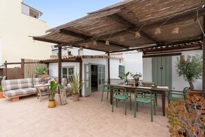 Een overdekt terras met een bank, eettafel, stoelen en potplanten.