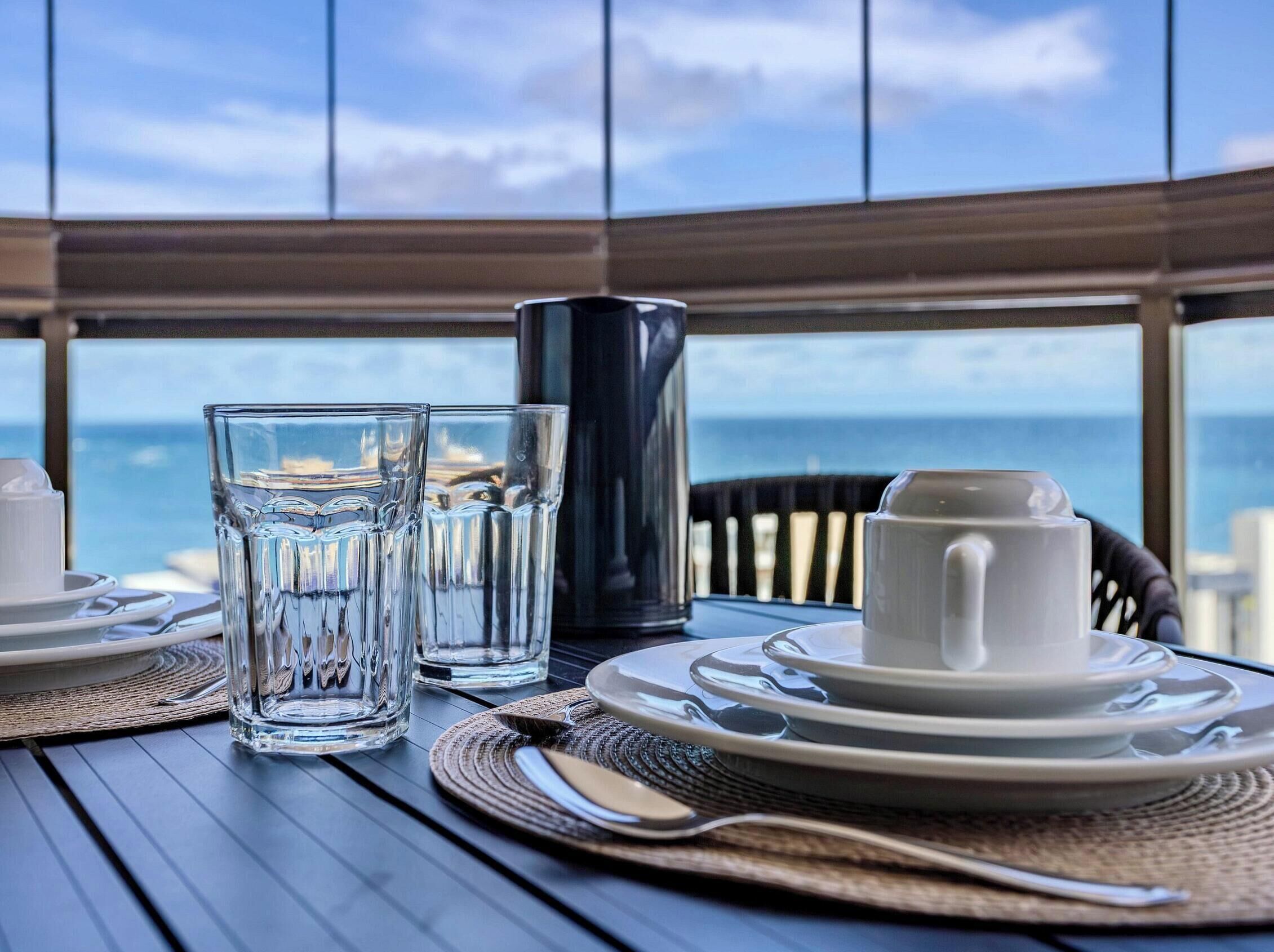 A table set with plates, glasses, and a cup, overlooking the ocean.