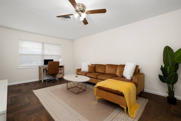 Ein Wohnzimmer mit braunem Sofa, Couchtisch, Schreibtisch mit Stuhl, Deckenventilator und einer großen Pflanze.