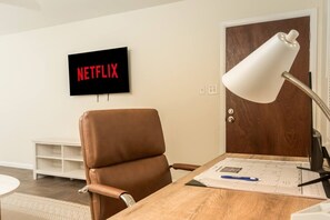 Et værelse med en brun læderstol, et træskrivebord med papirer og en pen, en hvid skrivebordslampe og et vægmonteret fjernsyn, der viser 'NETFLIX'.