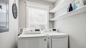 Une buanderie équipée de deux lave-linge et deux sèche-linge LG à chargement frontal, de couleur blanche, d’une fenêtre avec des stores et d’une étagère où sont rangées des bouteilles.