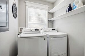 A laundry area with two white LG front-loading washers and dryers, a window with blinds, and a shelf with bottles. - Garden District Luxury Oasis-Private Garage (Baton Rouge)
