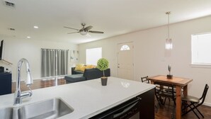 Ett modernt kök med diskmaskin, en matplats med ett träbord och stolar, en takfläkt och ett vardagsrum med en soffa och en växt.