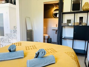 Quarto de hotel com cama, banheiro, espelho e prateleira com objetos decorativos.