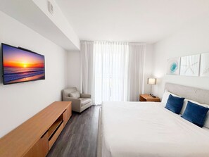 Habitación de hotel con cama, una silla, un escritorio, una mesita de noche, una lámpara y un cuadro de un atardecer.