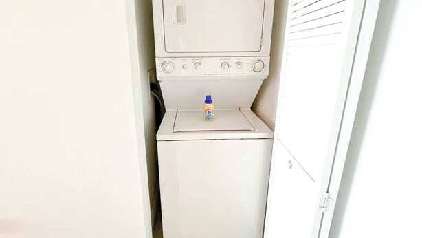 Una cambra de bugaderia petita amb una rentadora i una assecadora, una prestatgeria amb una ampolla de detergent, i una porta amb un pom.