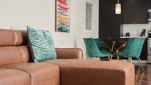 Un canapé en cuir marron avec un coussin vert, une chaise turquoise et, au fond, une cuisine moderne.