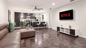 Un salon moderne comprenant un canapé d’angle, un coin repas, une cuisine et un téléviseur fixé au mur qui affiche « NETFLIX ».