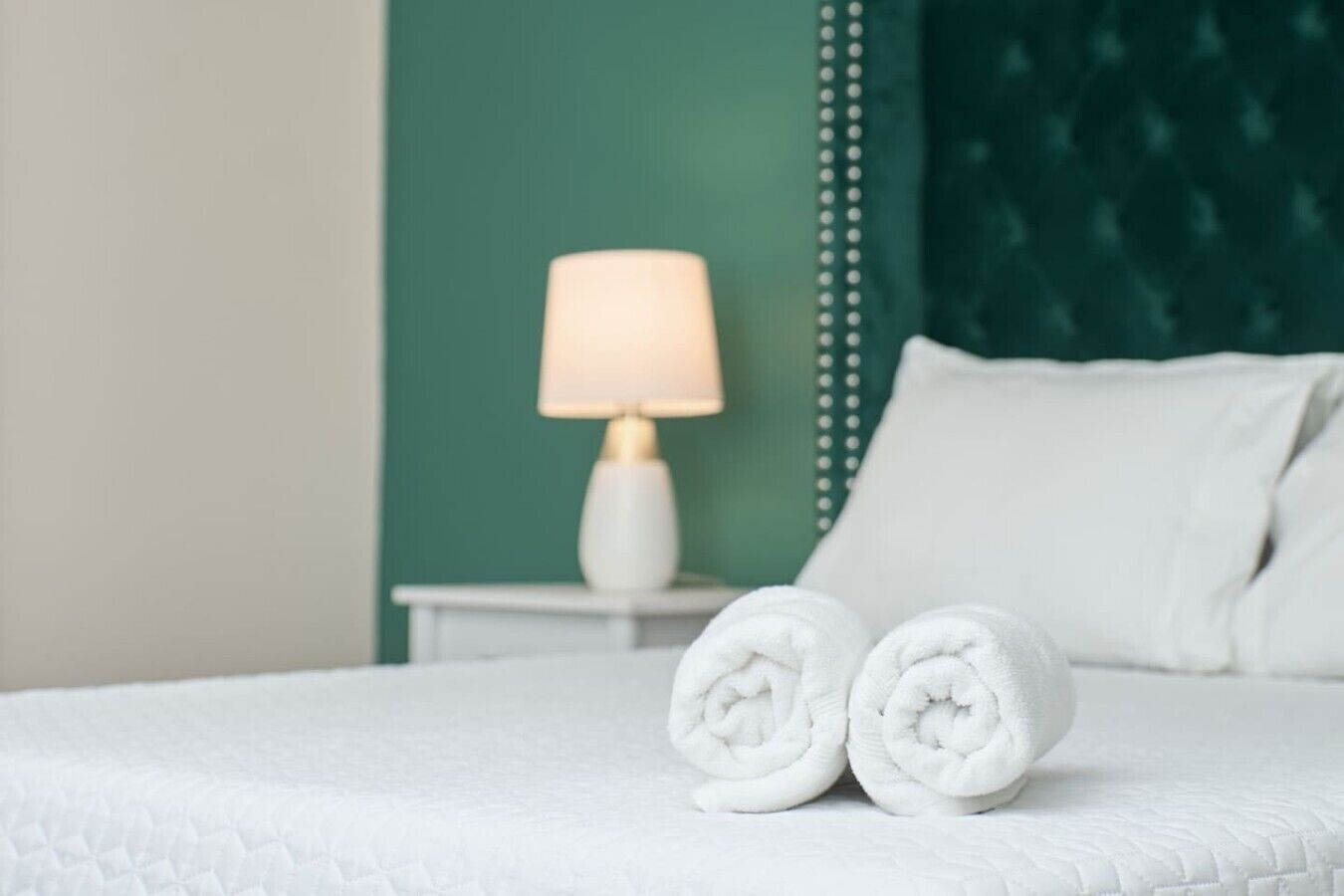 Un lit bien fait, avec du linge de lit blanc, une table de chevet blanche avec une lampe, et un mur d’accent vert.