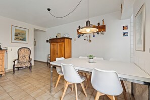A dining area with a white table, four white chairs, a wooden cabinet, and a hanging light fixture. - Westduin GV003 + G19* (De Panne)