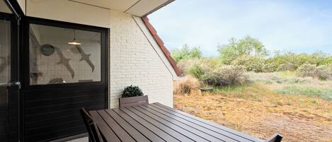 A patio with a wooden table and chairs, a black door, and a view of a dry landscape with bushes.