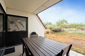 A patio with a wooden table and chairs, a black door, and a view of a dry landscape with bushes. - Westduin GV003 + G19* (De Panne)