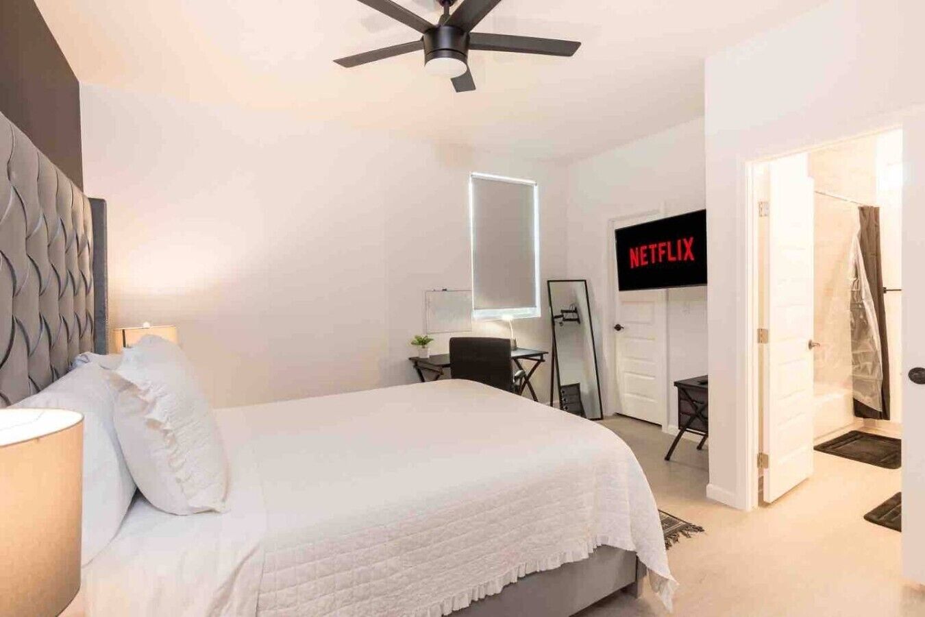 Ett sovrum med en säng, en takfläkt, en tv-skärm som visar ”NETFLIX”, en dörr och ett litet bord med en växt.