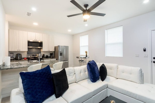 A modern living room with a white sectional sofa, a ceiling fan, and a kitchen in the background.