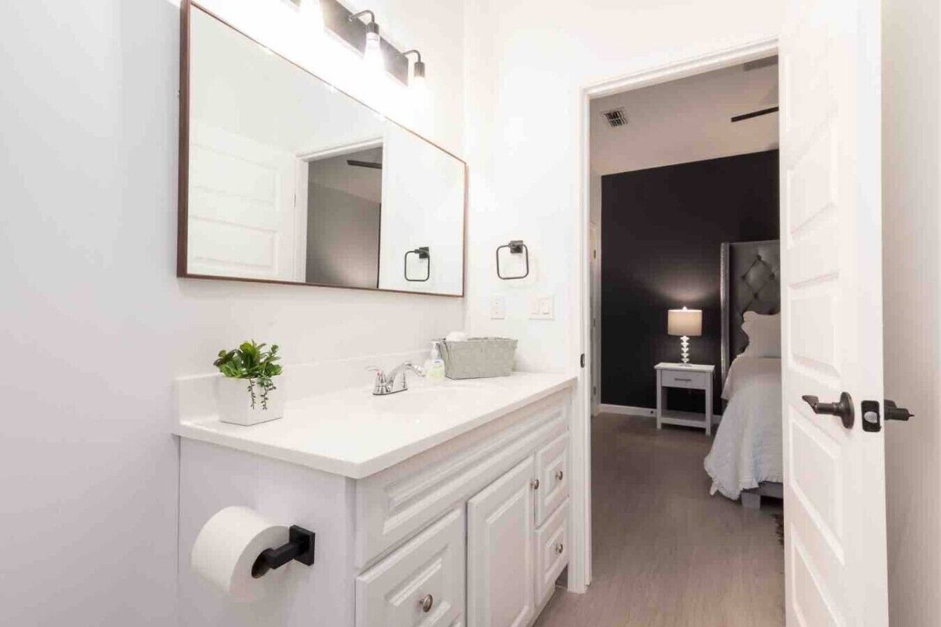 Un baño moderno con un espejo grande, un inodoro, un lavabo con una planta y una mesita de noche con una lámpara en el dormitorio contiguo.