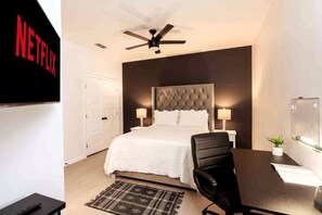 A hotel room with a bed, a desk, a chair, a ceiling fan, a TV, and a wall-mounted artwork.