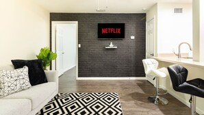 A modern living room with a dark brick wall, a flat-screen TV mounted above it, a ceiling fan, a white sofa with patterned cushions, a black and white rug, and a kitchen area with a sink and bar stools. - Lux, First Floor, Fast-WiFi King Size (McAllen)