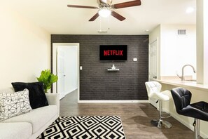 Ein modernes Wohnzimmer mit dunkler Ziegelwand, einem flachbildfernseher darüber, einem Deckenventilator, einem weißen Sofa mit gemusterten Kissen, einem schwarz-weißen Teppich und einer Küchenzeile mit Spüle und Barhocker.