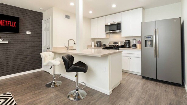 A modern kitchen with white cabinets, a stainless steel refrigerator, a microwave, and a coffee maker. There is a bar stool and a black chair in the dining area. - Lux, First Floor, Fast-WiFi King Size (McAllen)