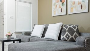 A grey sectional sofa with three pillows, a small black table with a vase, and a window with blinds. - King Size Beds/ Fast WiFi / Spacious/ Close to DHR (Edinburg)