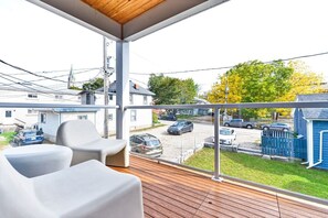 A balcony with two white chairs, a wooden deck, and a view of a street with cars and houses.