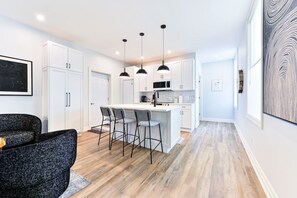A modern kitchen with white cabinets, a central island with bar stools, pendant lighting, and a wooden floor.