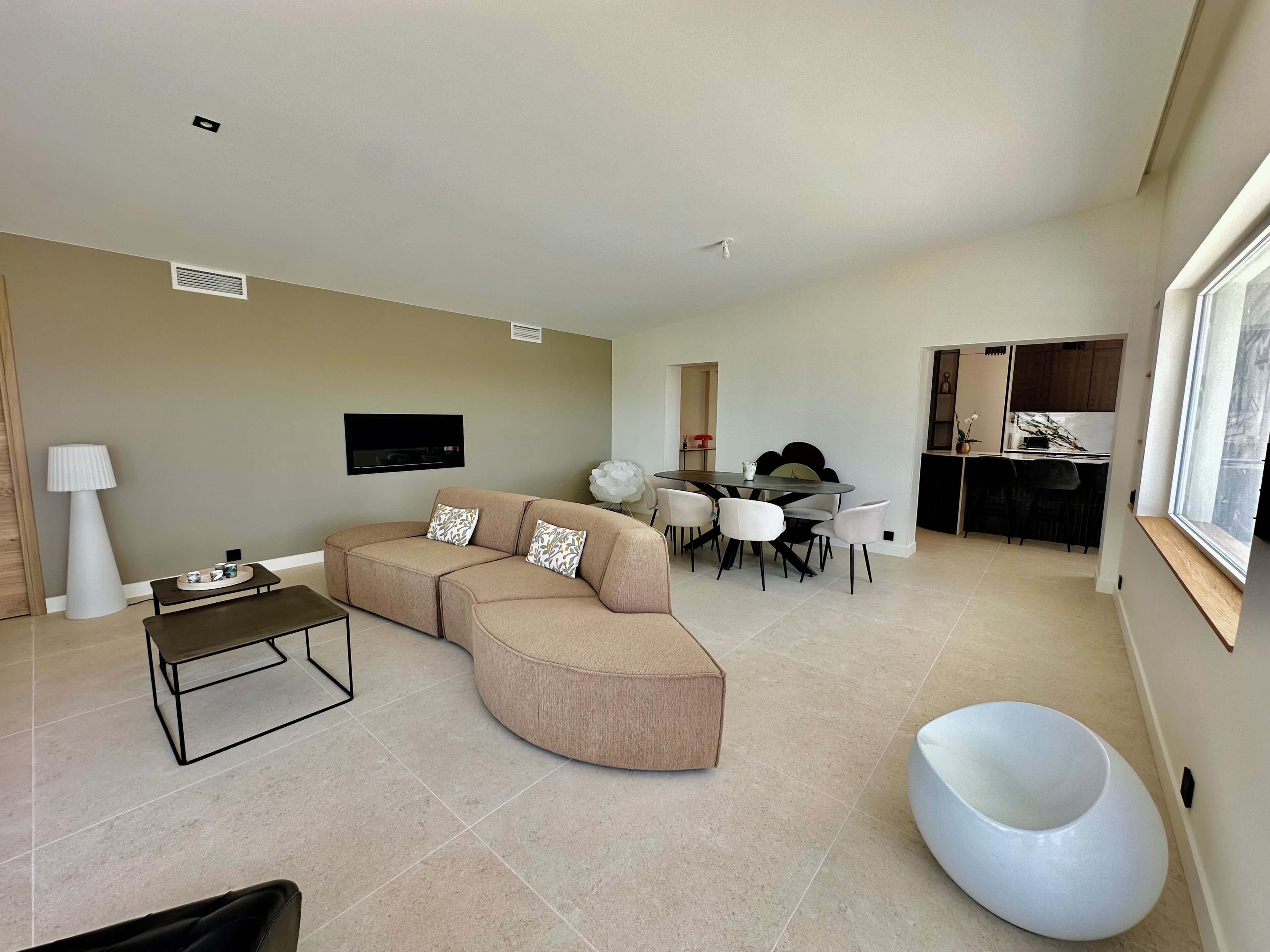 Ein modernes Wohnzimmer mit Ecksofa, einem Esstisch mit Stühlen, einem an der Wand befestigten Fernseher und einer Küchenzeile mit Schränken und einem Fenster.