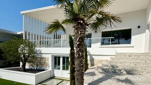 A modern building with a palm tree in a planter, a paved courtyard, and a white fence. - ROYAN PONTAILLAC UPSCALE VILLA "LES PINS" 2/4/6 PERSONS WITH POOL (Royan)
