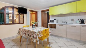 Een keuken met gele kastjes, een eettafel met een bloemig tafelkleed en een aan de muur bevestigde televisie.