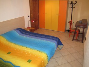 A bedroom with a bed, a wooden door, an orange wardrobe, a small table with a chair, and a coat rack.