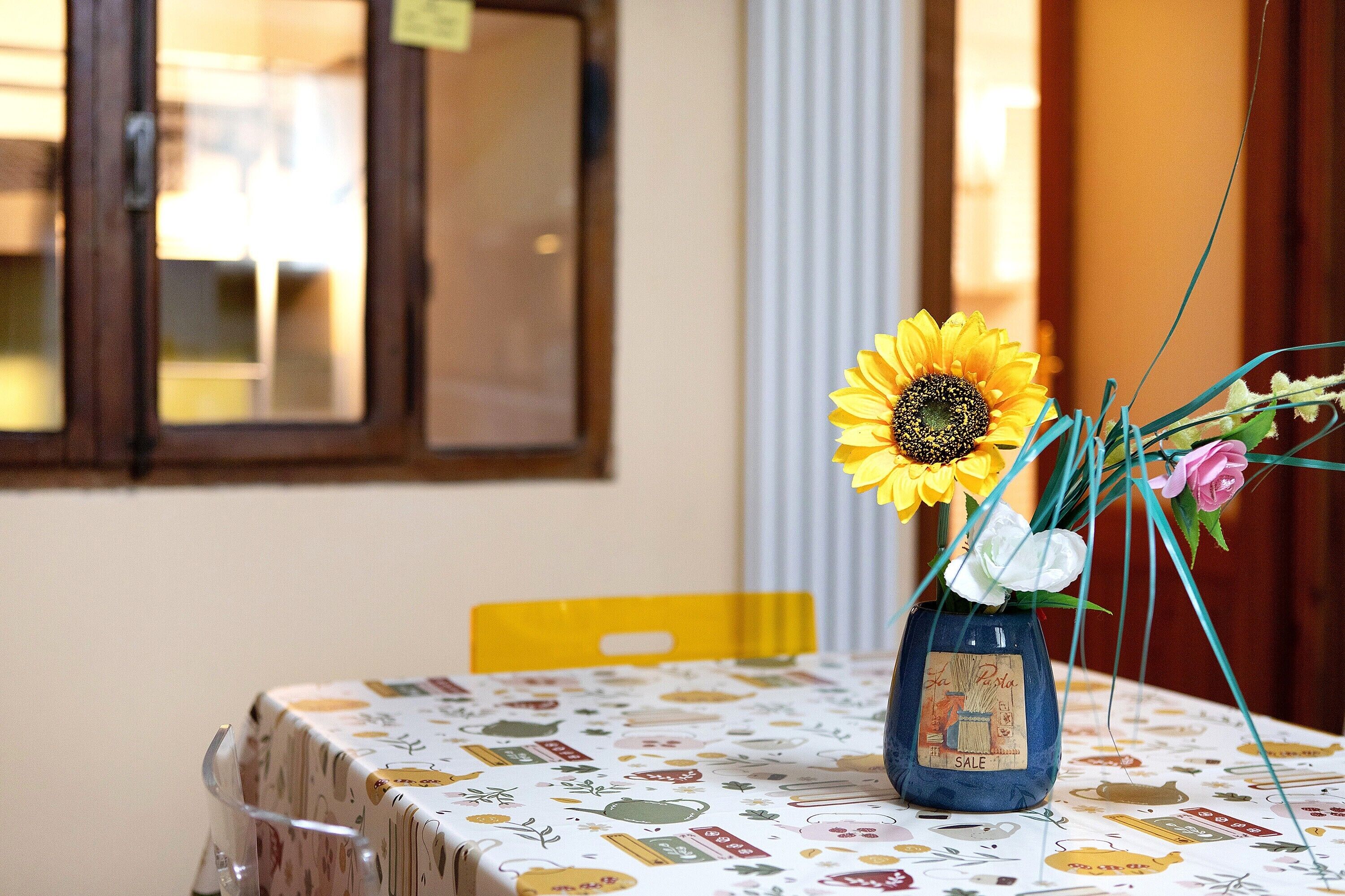 Une table recouverte d’une nappe à motifs, un vase rempli de fleurs et une fenêtre en arrière-plan.