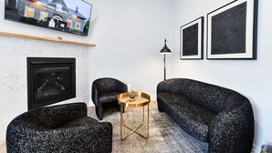 Una sala de estar moderna con chimenea, dos sillones negros, una mesa de centro redonda y un televisor montado en la pared.