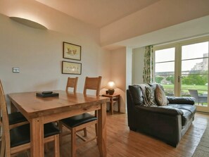 Un coin repas avec une table et des chaises en bois, un canapé et une lampe. Des tableaux encadrés ornent le mur, et une grande fenêtre offre une vue sur l’extérieur.