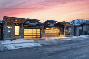 Une maison moderne à deux étages, dotée d’un garage, d’une façade en pierre et offrant une vue sur des montagnes enneigées.