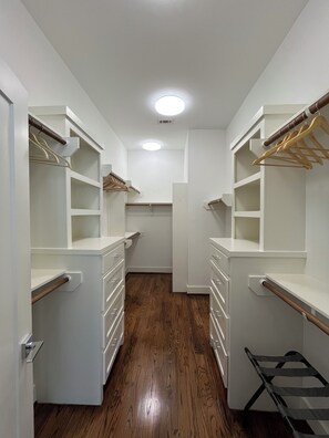 A narrow walk-through closet with white shelving units, wooden flooring, and a central light fixture.