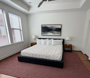 A bedroom with a bed, two bedside tables, a ceiling fan, a large window with blinds, and a wall-mounted painting.