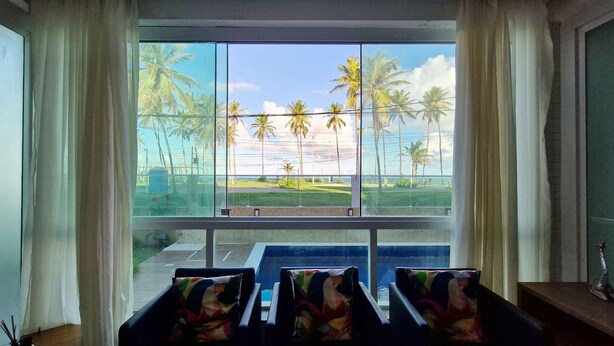 A room with a large window, a view of palm trees, and a small table with chairs. - COSTE0114 - Inba Stella Residence (Salvador)